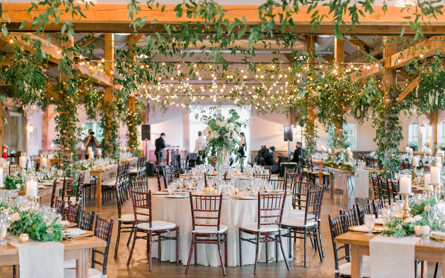 Event barn showcasing greenery, with white table rounds and greenery on tables along with floral arrangements.