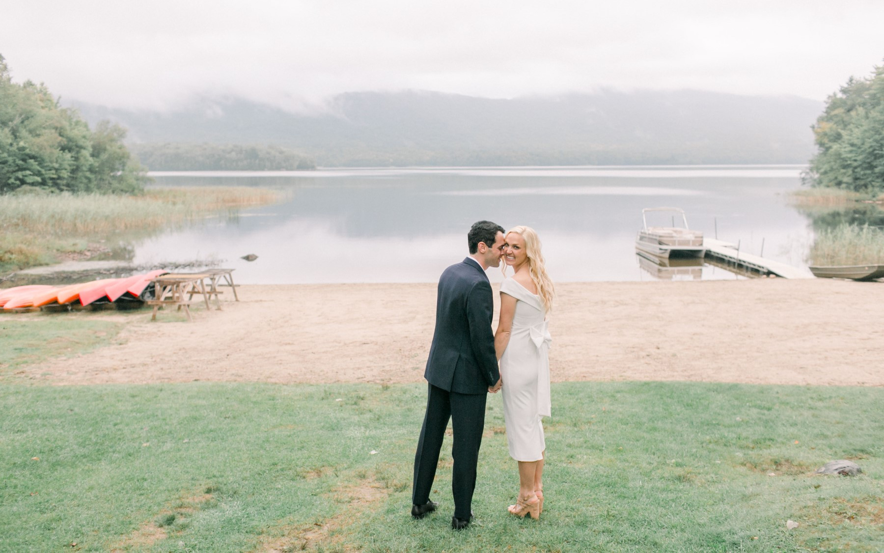 bride and groom on grass before beach, overlooking kayaks, lake, and pontoon boat