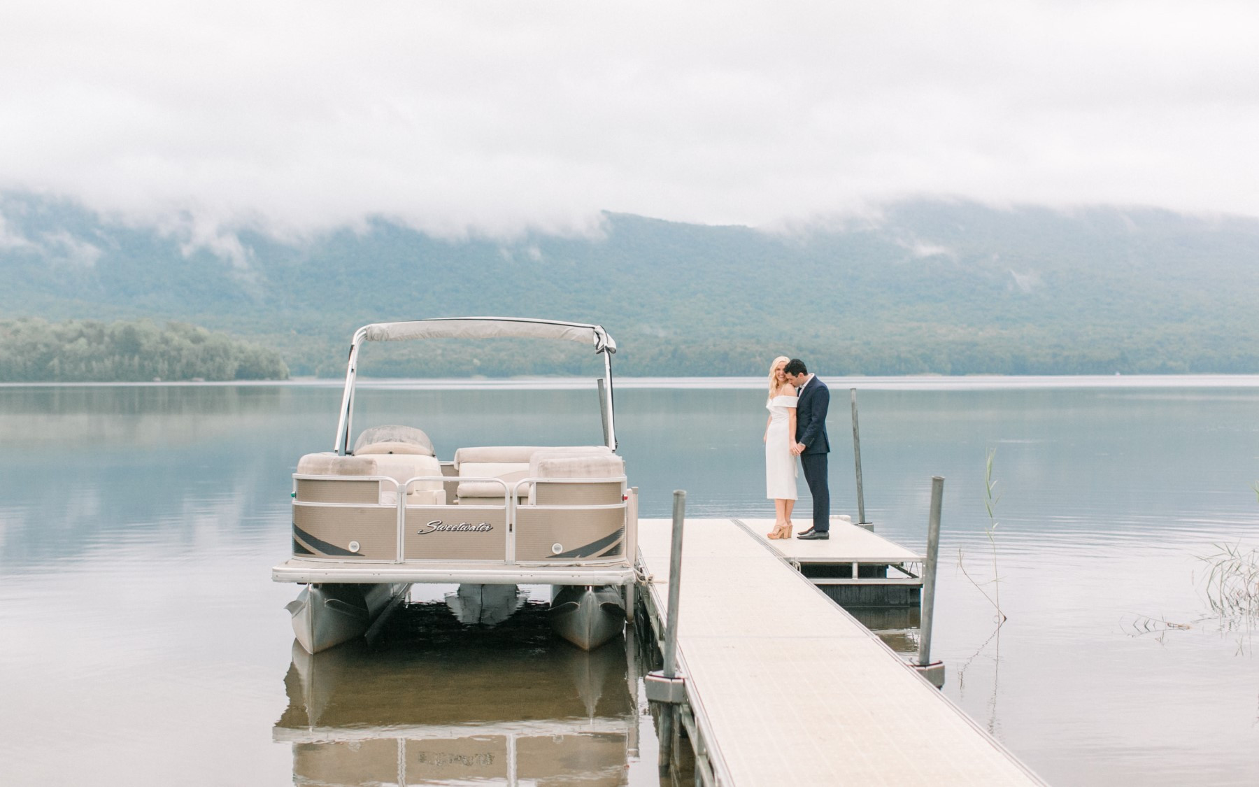 bride and groom on dock overlooking lake with pontoon boat to their left.