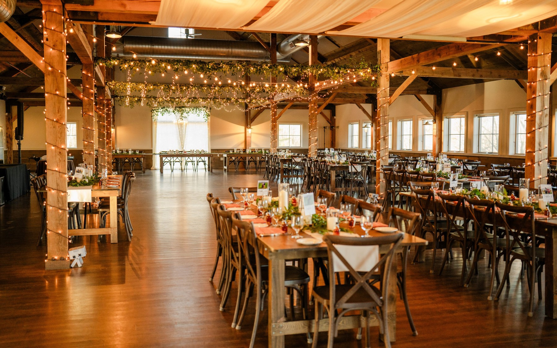 event barn filled with farmhouse tables and chivari chairs with greenery and drapery hanging from rafters.