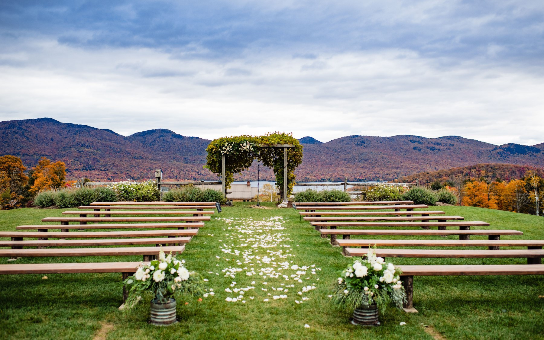 photo of fall wedding knoll with flowers at base of farmhouse benches and petals in alley with mountains and lake behind.