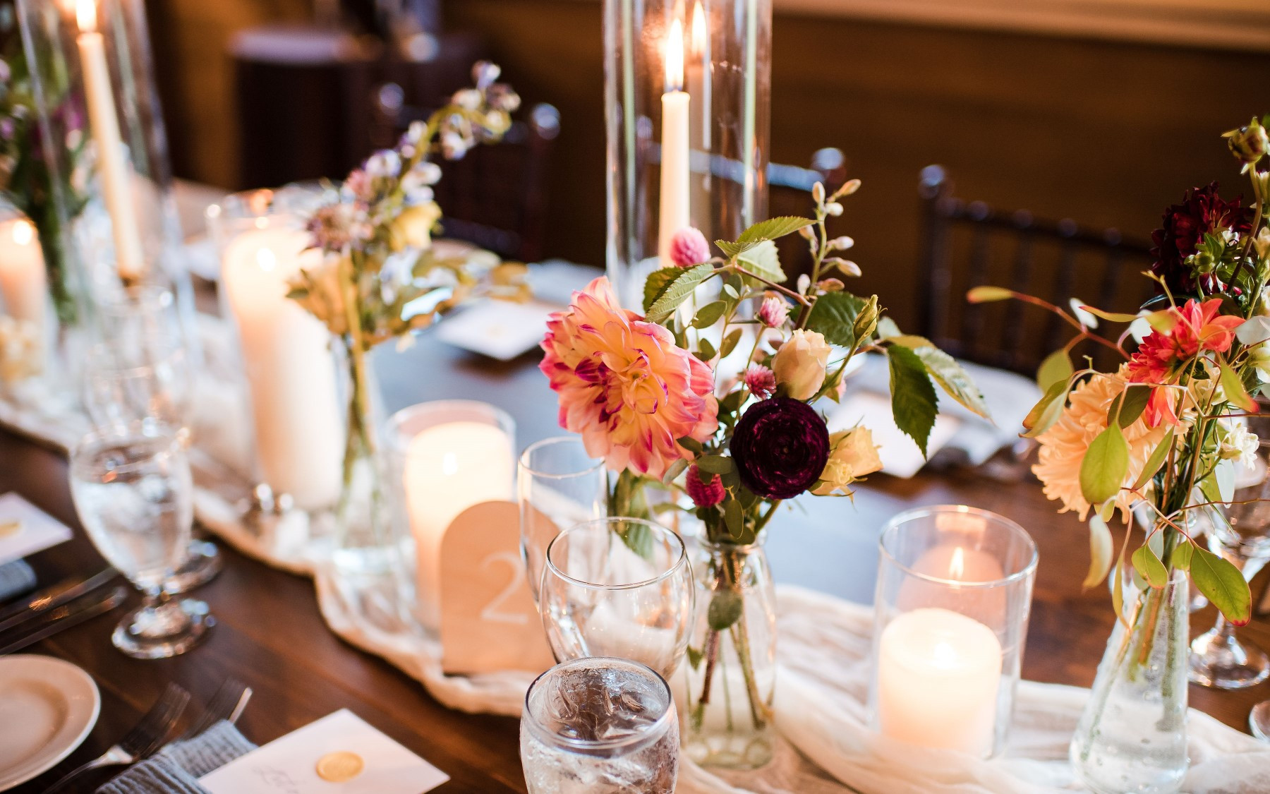 floral designs on farmhouse table featuring red, orange, blue, and green florals. Table is lined with candle votives and linen.
