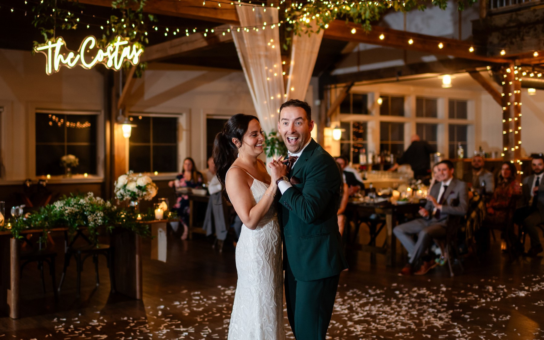 bride and groom in event barn holding hands while doing their first dance. drapery, lights, and greenery surround them with guests in the background in the event barn.