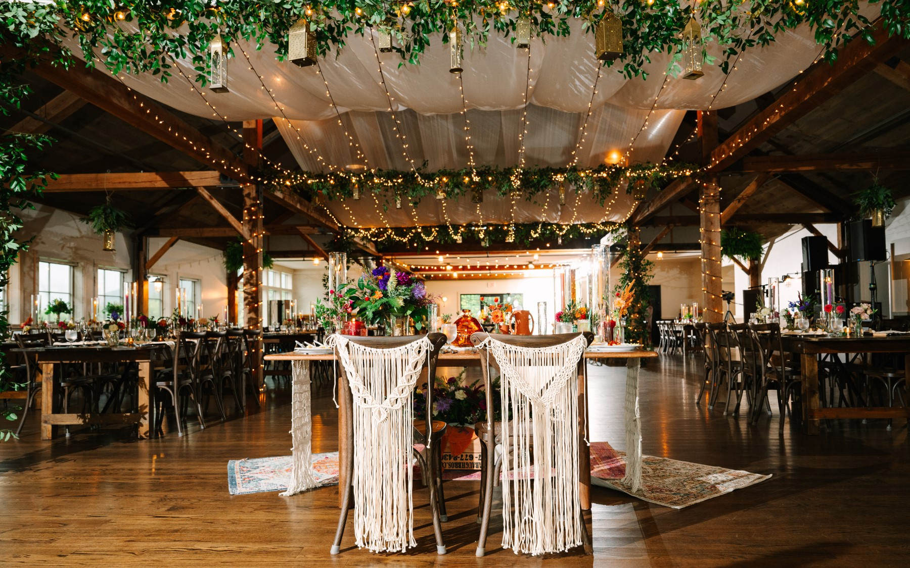 event barn decor for a boho style wedding, featuring overhead drapes, lighting, and greenery. sweetheart table is center of photograph with macrame designed backers for the chairs and floral is on the table.