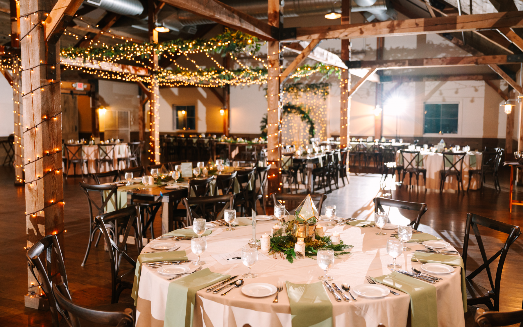 event barn set up with rounds and farmhouse tables with green napkins and greenery hanging from lights.