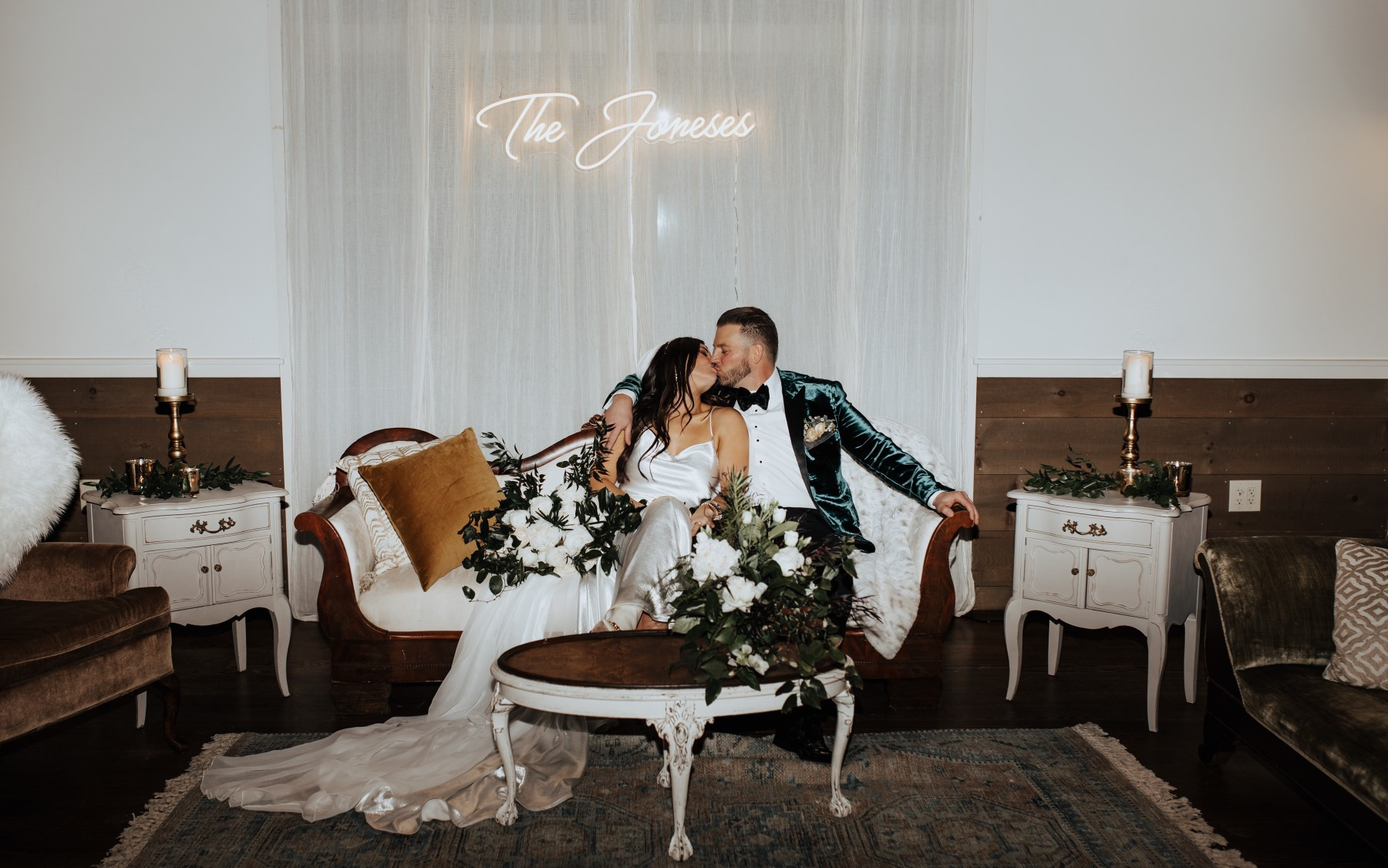 bride and groom sitting on velvet couch with floral arrangement kissing in front of sign that says \"The Joneses\"
