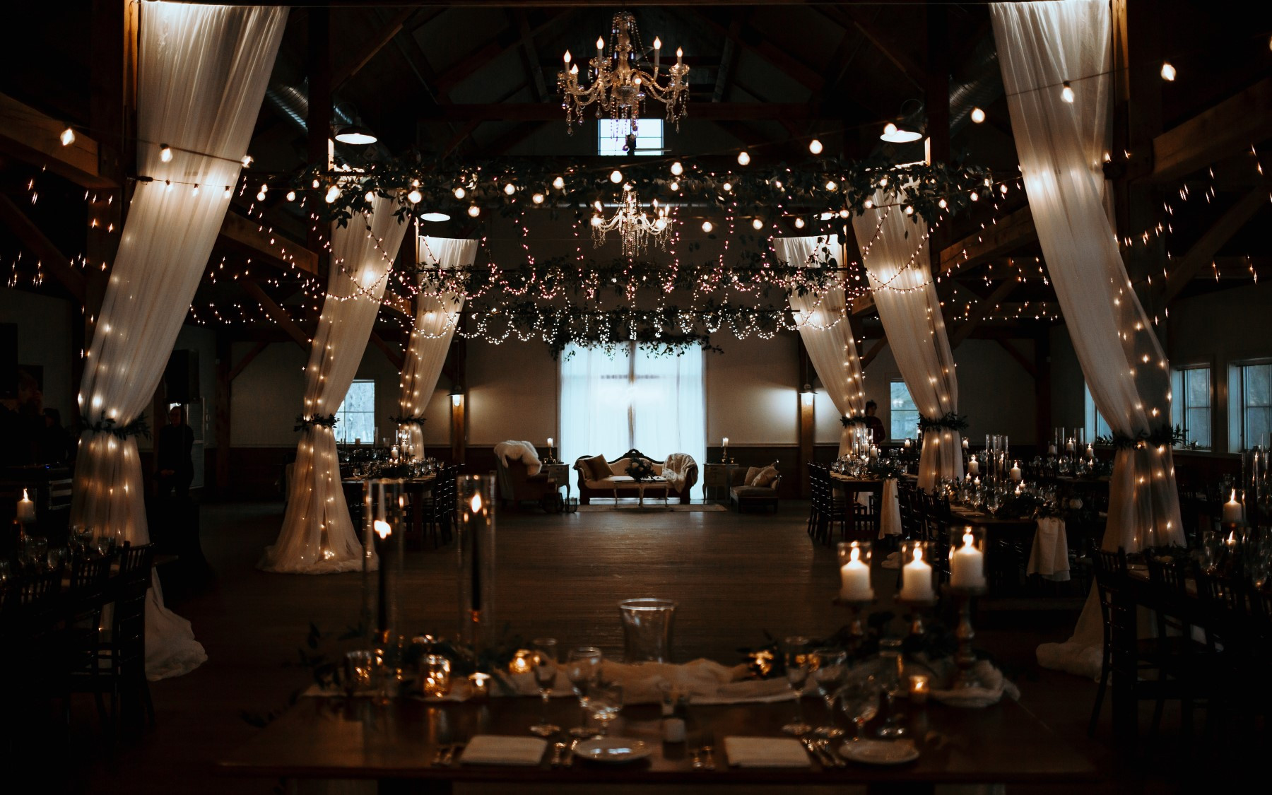 event barn covered in lighting, drapery, chandeliers, floral arrangements with seating at end in front of windows.