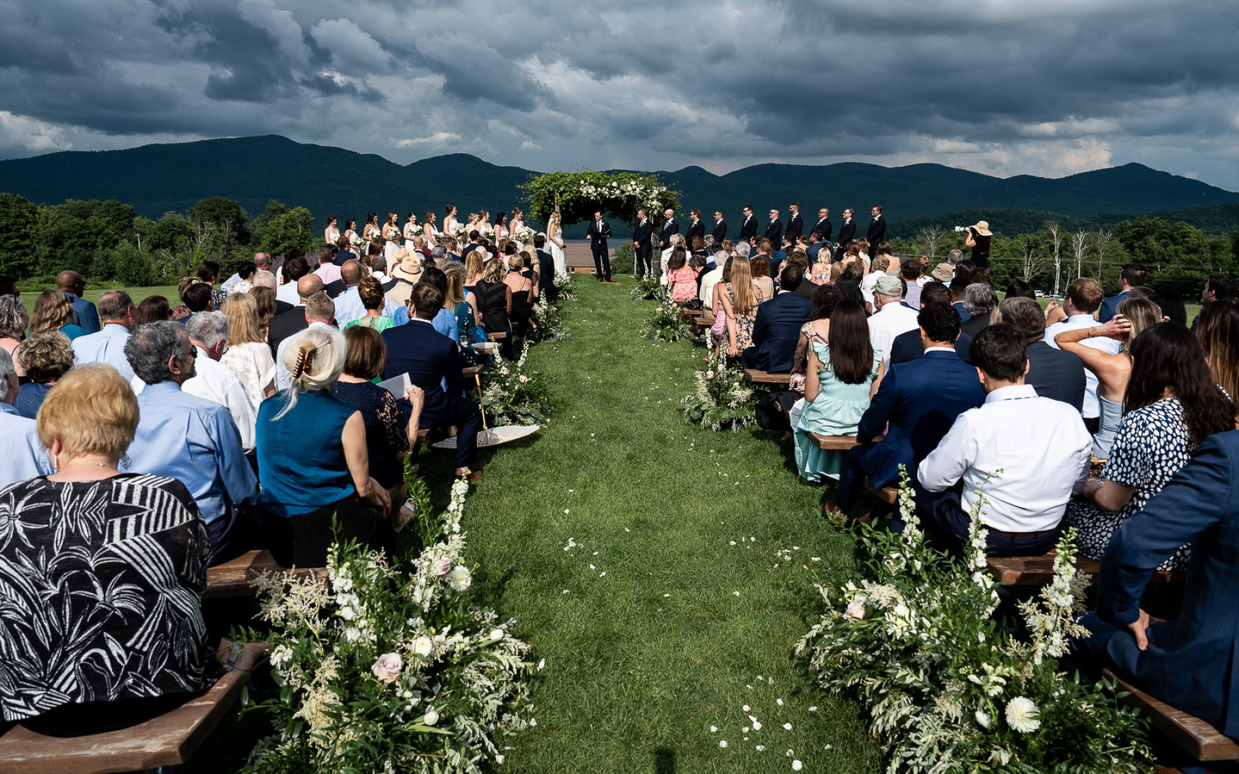 wedding knoll featuring guests sitting around on farmhouse benches with mountains in the background.