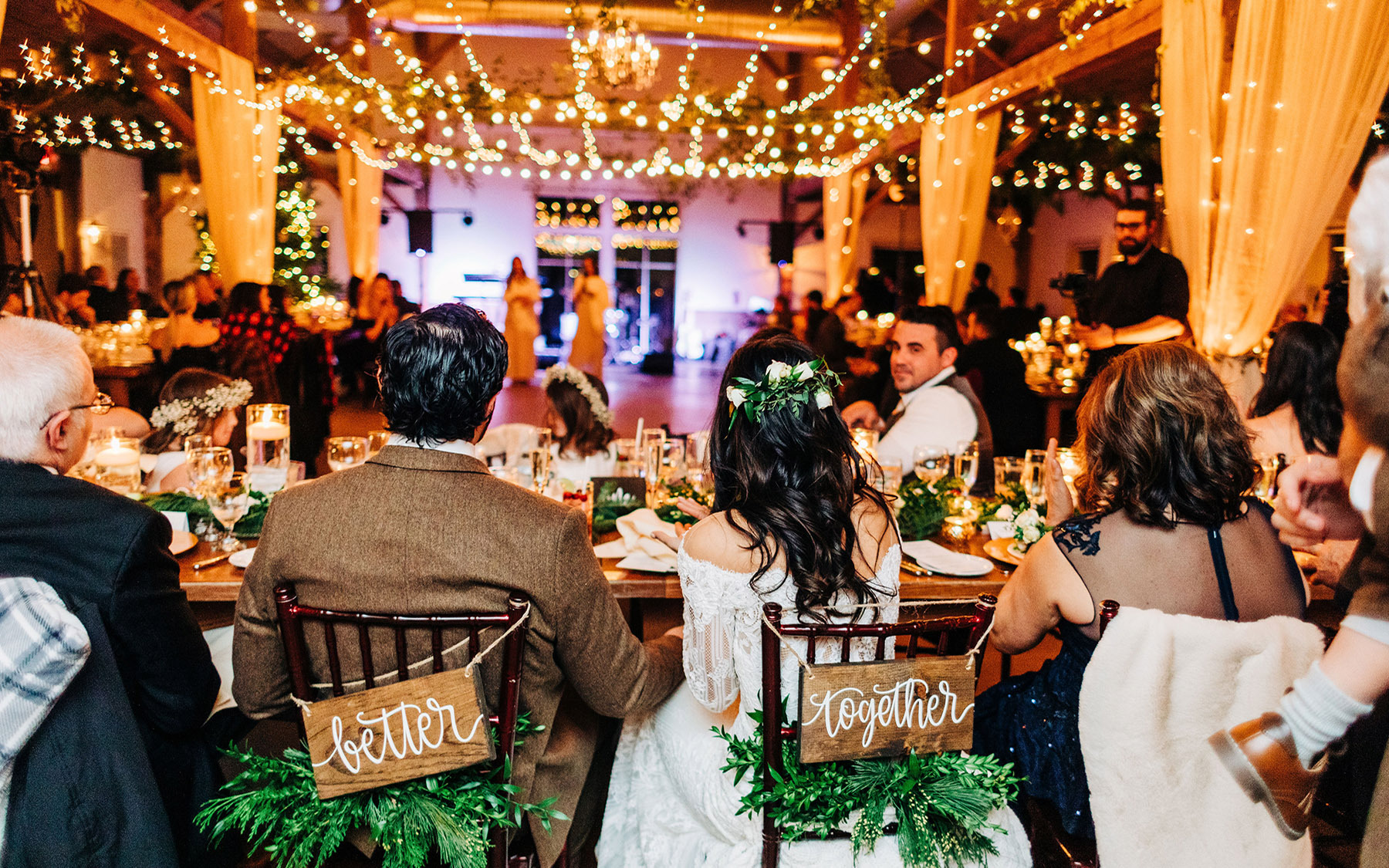 bride and groom sitting in chairs with their back to the camera looking at a room filled with people at tables and white lights and white drapery strung across the rafters.