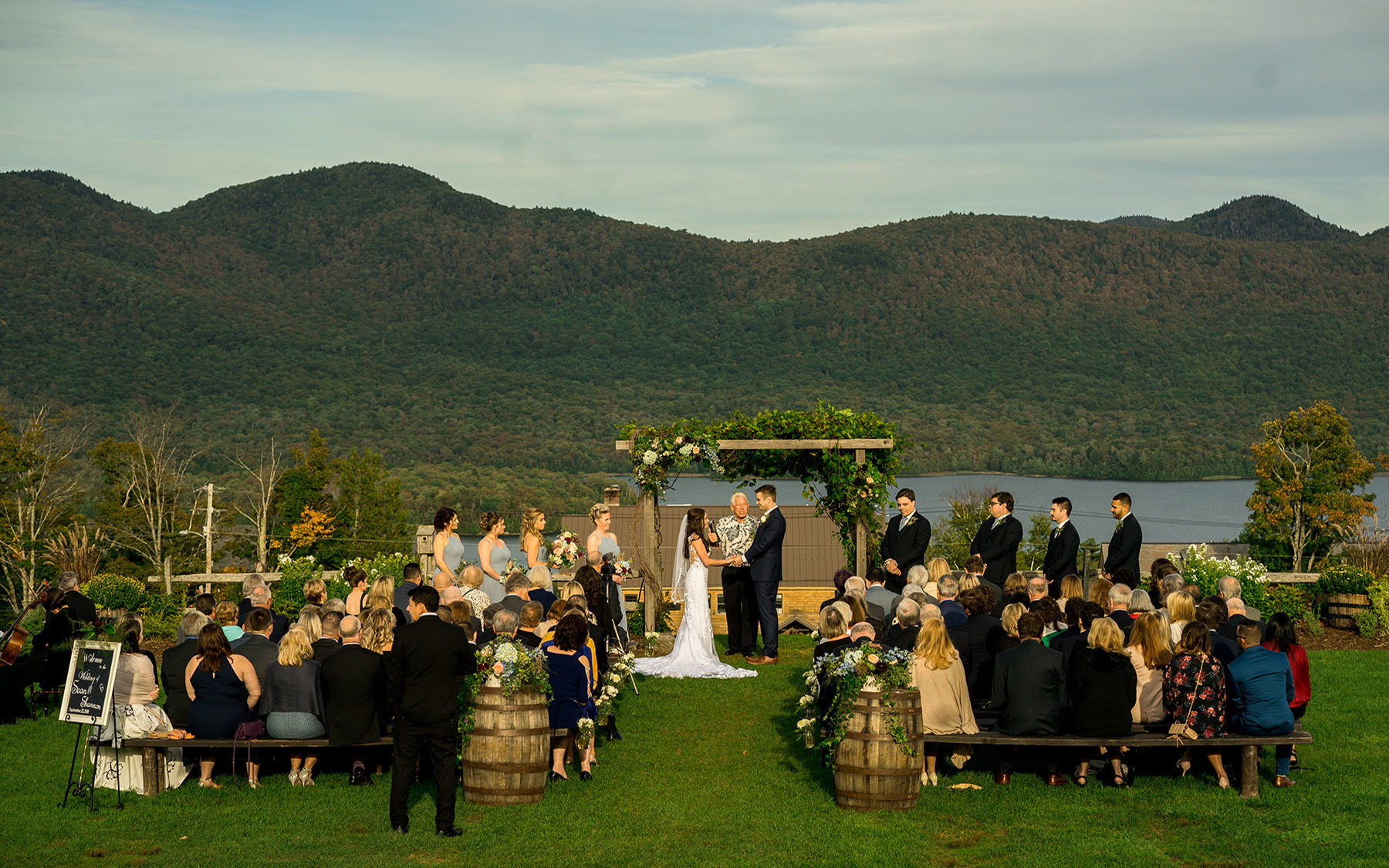 Group of people at outdoor wedding in various attire sitting on wooden benches in front of floral archway with bridal party and groom party in front overlooking lake and mountain at Mountain Top Inn & Resort in Chittenden, VT