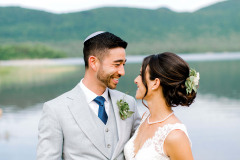 Brunette bride with hair up and brunette groom with grey suite standing in front of lake and green mountains.