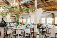 Room decorated for a wedding with white & blue hydrangea, white  tapered candles, white table clothes, greenery & string lights on the rafters, a chrystal chandelier and white draperies.