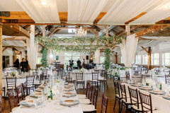 post and beam room decorated with white table clothes on tables with wooden chairs, white draperies, greens, a chandelier and string lights.