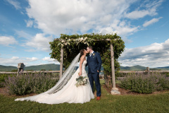 Bride in white, groom in dark suit, standing in front of arbor with greenery and white flowers.