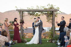 bride and groom in traditional dress with their arms around each other standing under an arbor kissing with a wedding party flanking their sides.