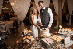 a wedding couple standing in front of a table full of desserts cutting a wedding cake. Drapery and tables visible in the background.