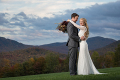 bride and groom in traditional dress with their arms around each other in a meadow with mountains and cloudy sky in the background
