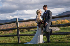 bride and groom in traditional dress with their arms around each other in front of a split rail fence with fall foliage and mountains in the background