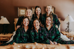 bride and bridesmaids sitting on a bed wearing green fleece pajama sets smiling at the camera. 