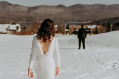 bride in white dress walking to groom in snowy meadow with lake and mountains in background. 