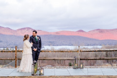 bride and groom walking on stone path. wooden fence behind them, and mountains in background. it is evening time, and the sun is setting, making the mountains look pink and purple. 