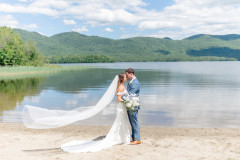 Groom and bride holding each other down by the lake's edge. The brides veil is flowing in the wind and the mountains are in the background.