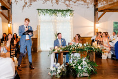 Bride and groom sitting at sweethart table in barn. Best man is giving a speech and all the guests surrounding are all laughing.