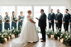 Loft floral ceremony site featuring green/white/blue floral arrangements, with bride and groom in front, and 3 groomsmen and 3 bridesmaids behind them
