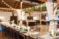 event barn featuring long farmhouse tables, greenery on tables with blue linens, and lights, greenery, and drapery from the top of the event barn.
