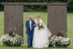 bride walking between farmhouse doors in arms with her father as they get ready to walk down the aisle.