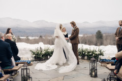 bride and groom holding hands exchanging rings while surrounded by guests on farmhouse benches. floral arrangement on ground behind them with mountainscape in background.