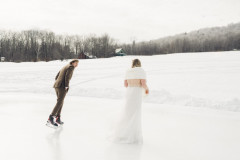 groom and bride skating on ice rink while in tuxedo and dress. background showcases a small red house and a small green house with mountains in the background.