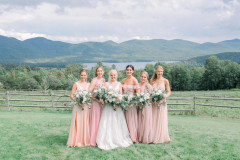 Bride in middle. 5 women in pink dresses, all carrying pink and white bouquets in front of lake and Green Mountains.