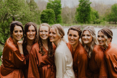 bride in white satin robe with six bridesmaids in red colored satin robes with pond and trees behind them.