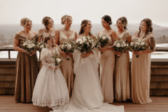 bride with bridal party standing in front of wooden deck fence with mountain scape in background smiling at each other. 