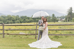 bride and groom standing under umbrella in front of wooden fence with mountains in background