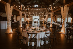 rustic event barn with lights hanging from rafters. there are circular and rectangular tables set up for a wedding reception with white linens.