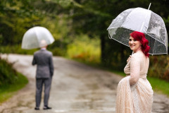 bride holding dress and umbrella with groom in distance, also holding an umbrella getting ready for the first look.