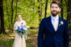 first look photo of bride holding floral bouquet in woods with groom in front