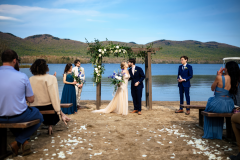 summer wedding with arbor with floral designs on it featuring bride and groom kissing underneath with green mountains and lake in background.