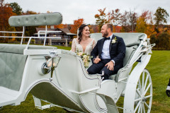 bride and groom in dress and suit sitting in a white carriage with fall foliage in background