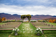 photo of fall wedding knoll with flowers at base of farmhouse benches and petals in alley with mountains and lake behind.