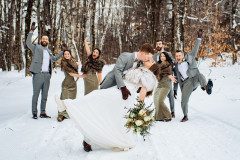 Bride and groom kissing with floral bouquet with groomsmen and bridesmaids in back in winter scape.