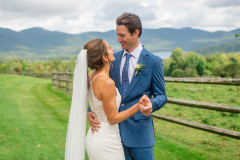 bride and groom smiling at each other holding hands in front of a rustic wooden fence with a green mountain landscape in the background.