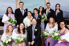 wedding party posing for camera. bride and bride are in the center. there are four members of the wedding party in black suits with purple ties, and five members in purple dresses and white shawls. 