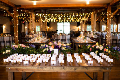 event barn with multi colored arrangements on farmhouse table and name cards with tables set for event in back with lights strung from rafters.