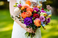 floral bouquet featuring red, orange, yellow, purple, blue, and white florals being held by bride.
