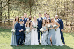 bride and groom with floral bouquet surrounded by 6 groomsmen and 6 bridesmaids all with white floral bouquets standing together in front of trees