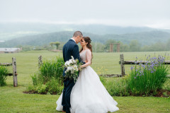 bride and groom standing in front of wooden fence overlooking wedding knoll and lodge with fog in front of mountains.