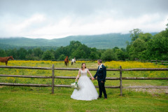 bride and groom standing in front of wooden fence. behind the fence is a horse pasture featuring two brown horses and one white and brown spotted horse. background includes mountains and fog.